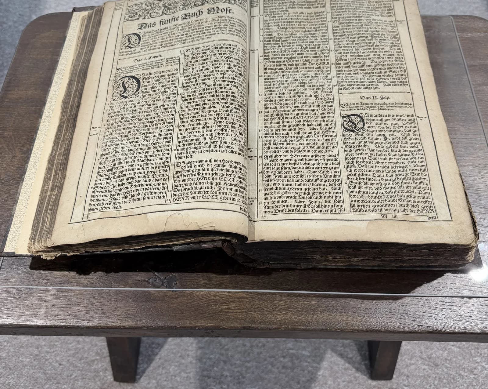 Duży starodruk Biblii otwarty na karcie z nagłówkiem „Das fünffte Buch Mose”, czyli Piątej Księdze Mojżeszowej, na drewnianym pulpicie w gablocie.