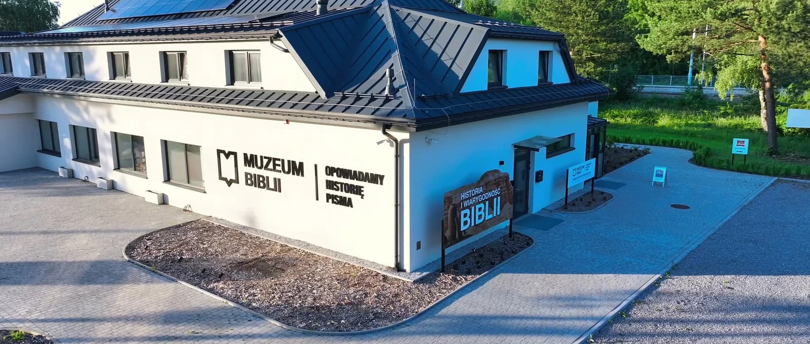 Biały budynek Muzeum Biblii z czarnym znakiem na ścianie, przed nim wyłożony kostką parking i wejście z tablicą „Historia i wiarygodność Biblii”.